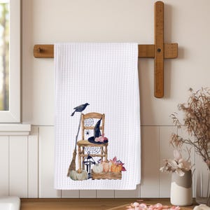 May include: A white waffle-weave kitchen towel featuring a watercolor illustration of a wooden chair, a black crow, a broom, a witch hat with pink flowers, spider webs, a lantern, and a wooden crate filled with assorted pumpkins and leaves.
