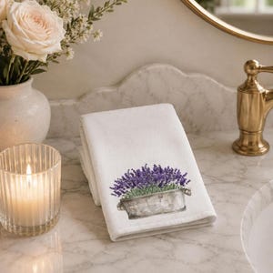 A white folded hand towel with a printed image of purple lavender flowers in a gray planter, on a marble countertop with a lit candle and a vase of white roses.