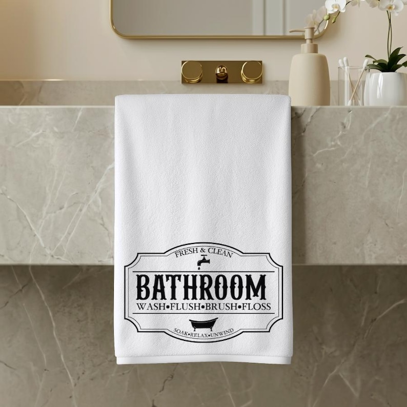 May include: A white hand towel featuring a black graphic design with the text "FRESH & CLEAN", "BATHROOM", and "WASH&bull;FLUSH&bull;BRUSH&bull;FLOSS" along with a small faucet icon and a bathtub icon centered within a decorative border frame.