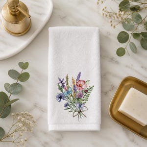 May include: A white hand towel featuring a printed watercolor illustration of a wildflower bouquet. The bouquet includes purple, pink, and blue flowers with green foliage and a small brown bow tied at the base of the stems, centered on the fabric.