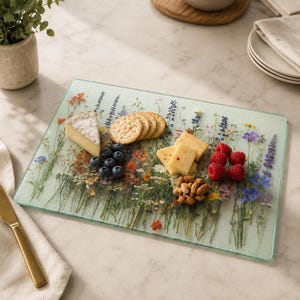 A glass cutting board decorated with a pressed floral pattern is used as a charcuterie platter, holding cheese, crackers, berries, and almonds on a marble surface.