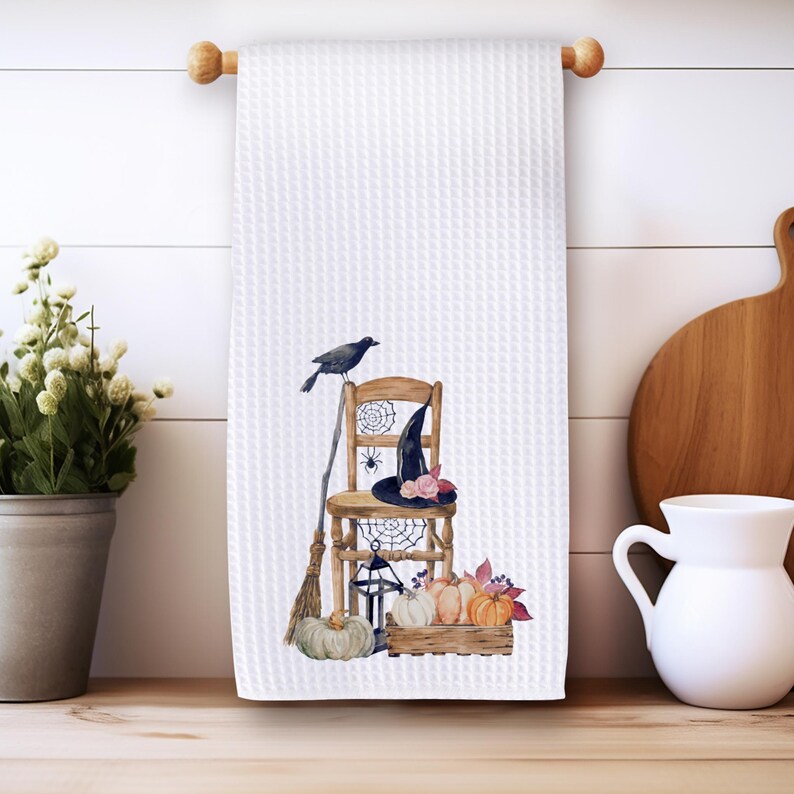 May include: A white waffle-weave kitchen towel featuring a printed illustration of a wooden chair, a black witch hat, a crow, a broom, a lantern, spiderwebs, and a wooden crate filled with orange and white pumpkins and autumn leaves.