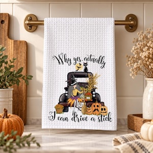 May include: A white waffle-weave kitchen towel featuring a graphic of a black truck, a witch, a black cat, and pumpkins. The text reads, "Why yes, actually I can drive a stick." The design includes orange, yellow, and black colors on the fabric.