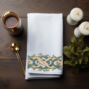 Może przedstawiać: Biały ręcznik dekoracyjny z zielonym, beżowym i niebieskim przeplatanym wzorem wzdłuż dolnej krawędzi. Złota łyżka i kubek ze złotym obramowaniem znajdują się po lewej stronie. Dwa białe świece i zieleń po prawej.
