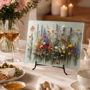 A glass cutting board decorated with pressed wildflowers is displayed upright on a dining table set with wine glasses, cheese, and a teapot.