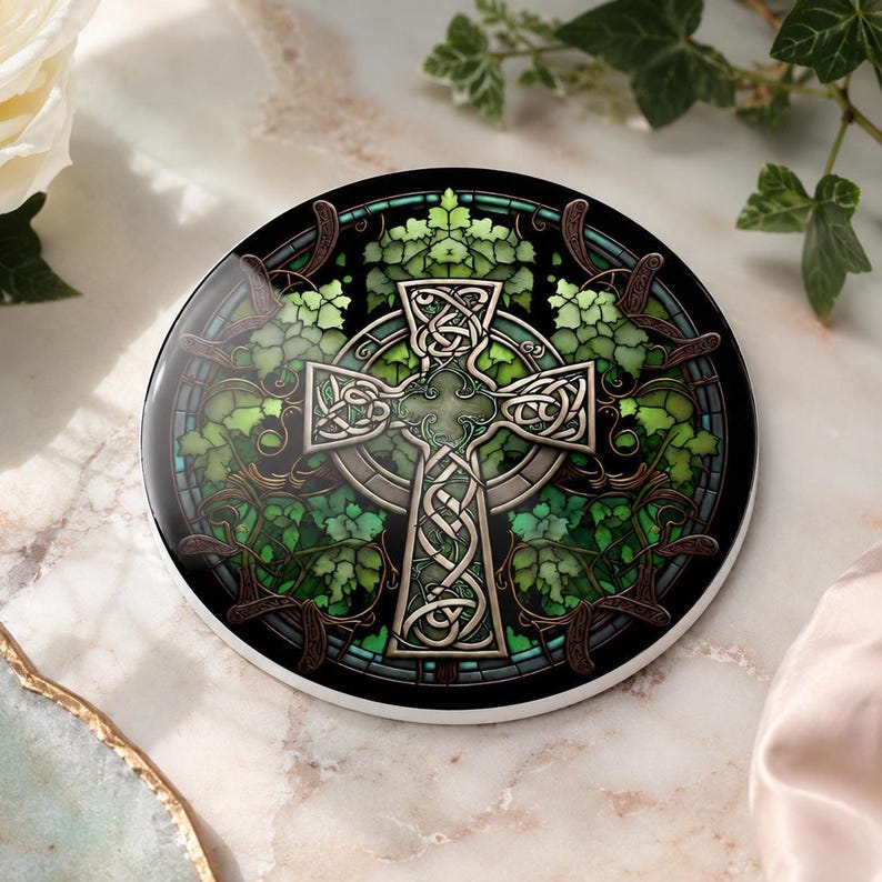 A round ceramic coaster with a Celtic cross design in shades of green and brown, surrounded by ivy-like foliage, on a marble surface with a white rose and green leaves.