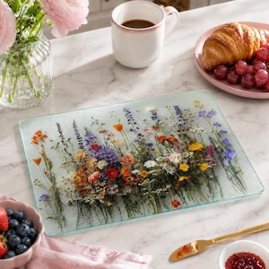 A glass cutting board featuring a pressed floral design sits on a marble table next to a croissant, bowl of berries, coffee mug, and jam.
