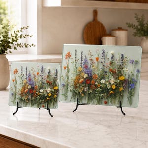 Two glass cutting boards of different sizes, both featuring the same colorful pressed wildflower design, stand on black display stands on a kitchen island.
