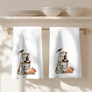 May include: Two white waffle-weave kitchen towels hanging on a gold rod. Each towel features a printed illustration of a wooden chair, a black witch hat, a broom, a crow, a spider web, a lantern, and a crate filled with pumpkins and autumn leaves.