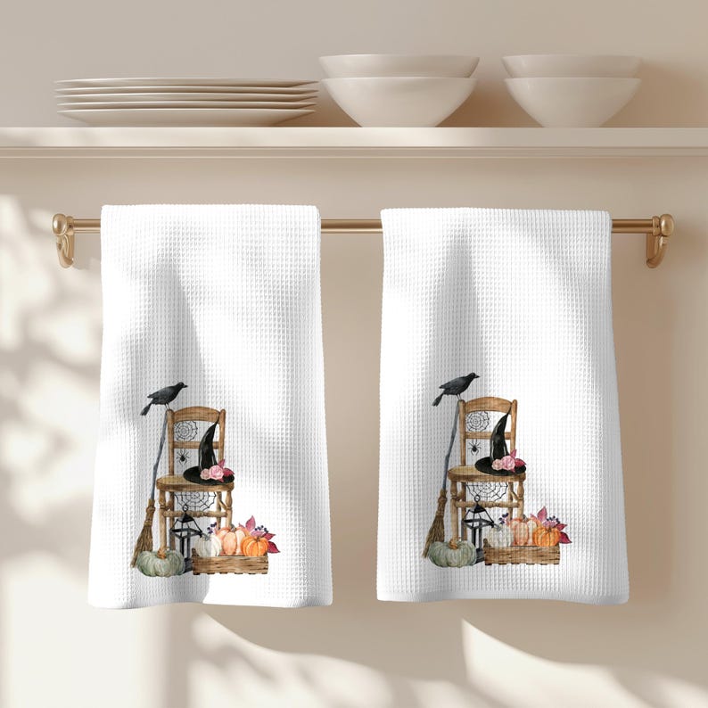 May include: Two white waffle-weave kitchen towels hanging on a gold rod. Each towel features a printed illustration of a wooden chair, a black witch hat, a broom, a crow, a spider web, a lantern, and a crate filled with pumpkins and autumn leaves.