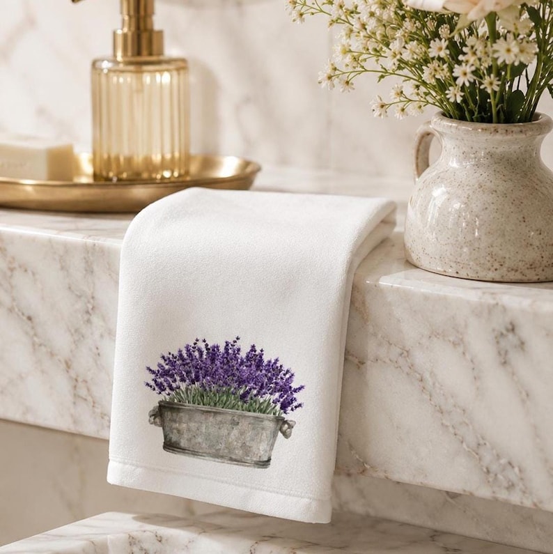 A white folded hand towel with a printed image of purple lavender flowers in a gray planter, resting on the edge of a white sink with a gold soap dispenser and a vase of white flowers in the background.
