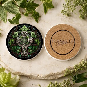 Two ceramic coasters, one with a green Celtic cross and ivy design, and the other showing a cork bottom with "FERNVILLE Creations" text, on a light stone surface with green plants.