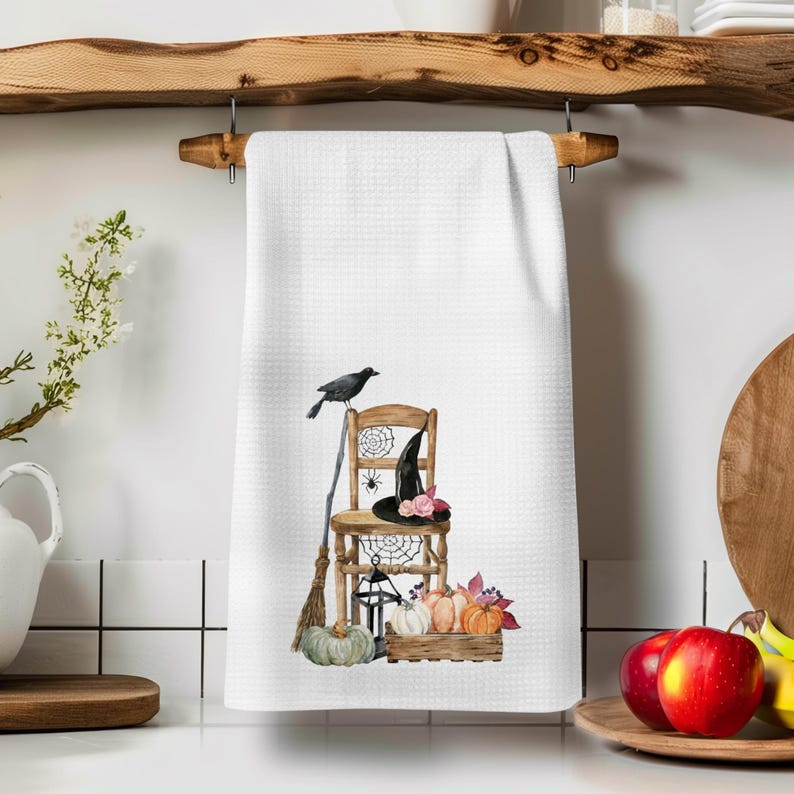 May include: A white waffle-weave kitchen towel featuring a printed illustration of a wooden chair, a black witch hat with pink flowers, a black crow, a broom, a lantern, spiderwebs, and a wooden crate filled with assorted pumpkins.