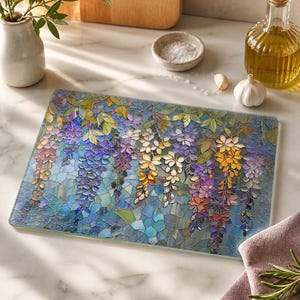 A rectangular glass cutting board with a stained-glass wisteria design in blues, purples, and greens rests on a white marble counter next to salt, garlic, and olive oil.