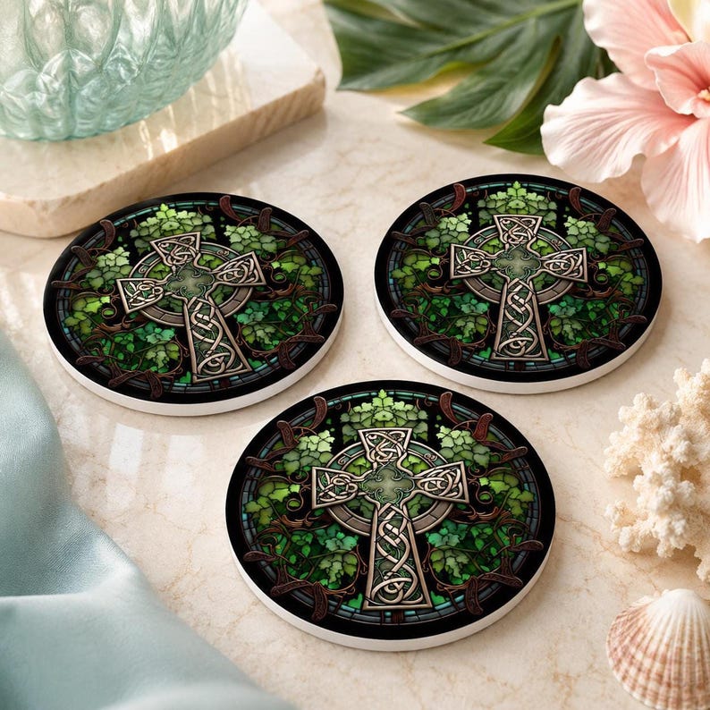 Three round ceramic coasters with a green Celtic cross and ivy design, arranged on a light blue and white marble surface with a pink flower, a teal glass, and white coral.