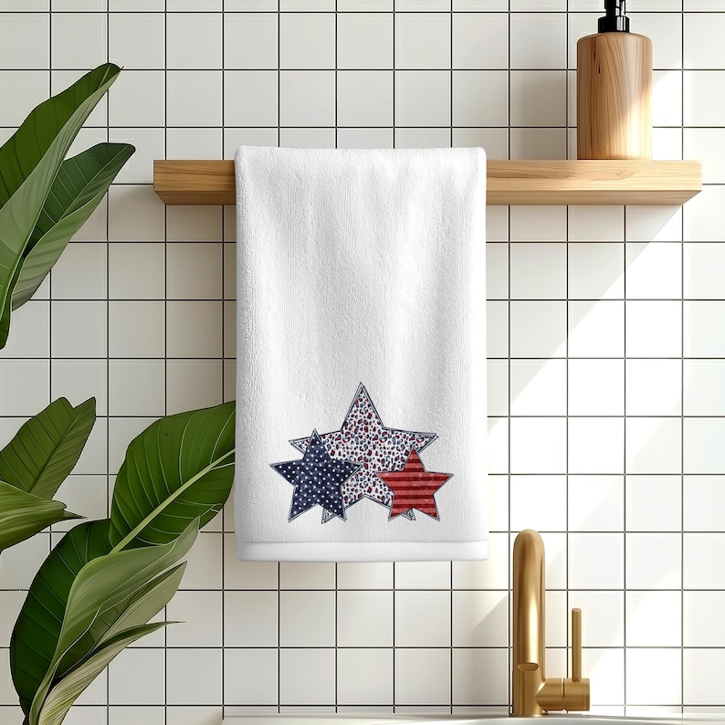 May include: White hand towel with three star designs in red, white, and blue. The stars have different patterns, including stripes and polka dots. The towel is hanging on a wooden rack in a bathroom setting.