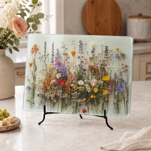 A glass cutting board with a pressed wildflower pattern is displayed on a kitchen counter next to a vase of flowers and a small plate of cheese and grapes.