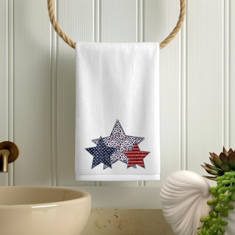 May include: White hand towel with three star designs in red, white, and blue. The stars have different patterns: polka dots, stripes, and a speckled design. The towel is hanging on a rope ring.