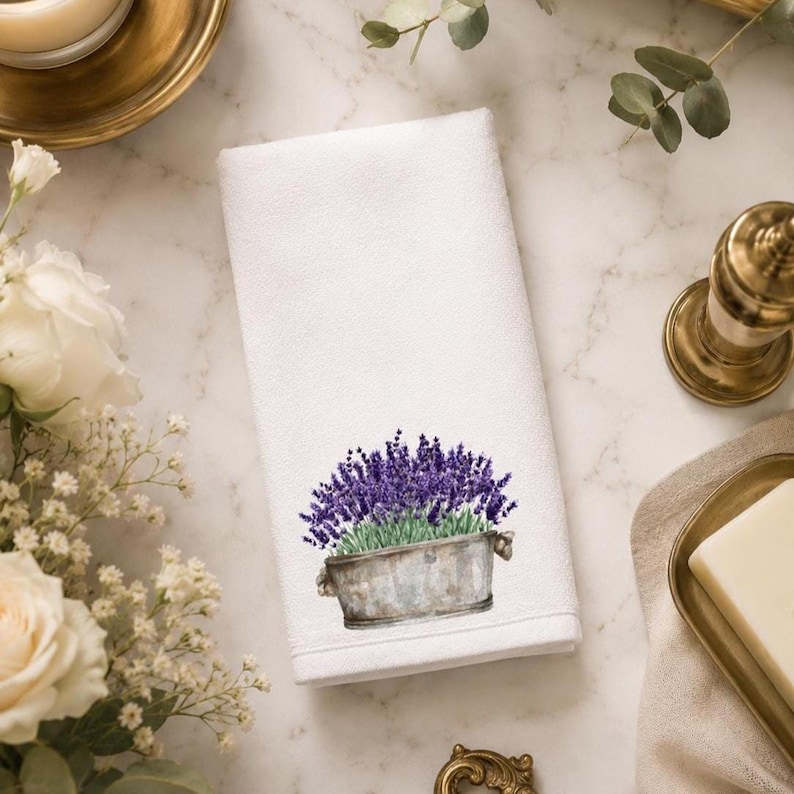 A white folded hand towel with a printed image of purple lavender flowers in a gray planter with "FLOWER" on it, on a marble surface with white roses and brass decor.