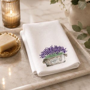 A white folded hand towel with a printed image of purple lavender flowers in a gray planter with "FLOWER" on it, on a light-colored tray with a lit candle and a bar of soap.