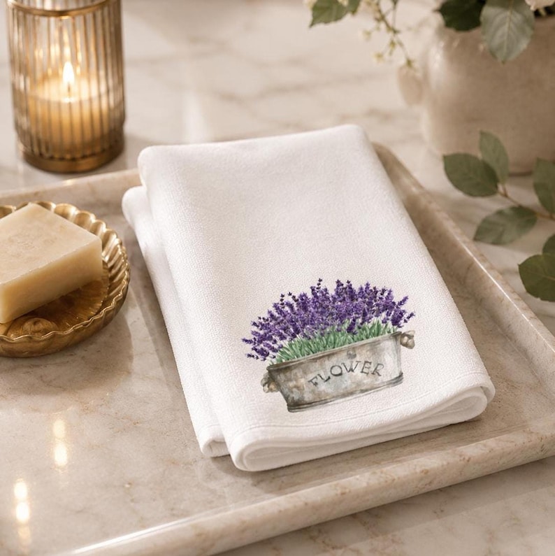 A white folded hand towel with a printed image of purple lavender flowers in a gray planter with "FLOWER" on it, on a light-colored tray with a lit candle and a bar of soap.