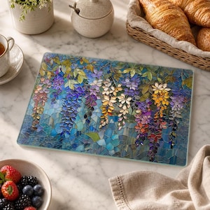 A rectangular glass cutting board with a stained-glass wisteria design is on a white marble counter, with a bowl of berries and a basket of croissants nearby.