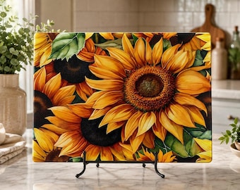 May include: A rectangular glass cutting board featuring a vibrant sunflower pattern with large yellow petals, dark brown centers, and green leaves. The board is displayed upright on a black metal stand, showcasing a detailed floral design.
