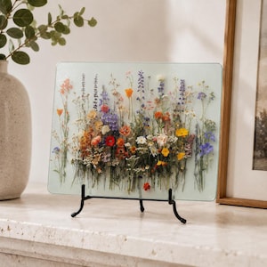 A rectangular glass cutting board with colorful pressed flowers stands upright on a black display easel on a stone mantle.