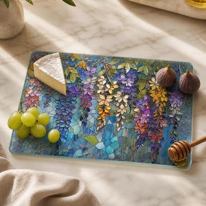 A rectangular glass cutting board with a stained-glass wisteria design holds a wedge of brie, green grapes, and two figs on a white marble counter.