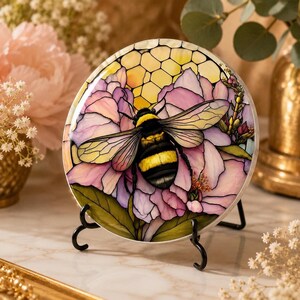 A round ceramic coaster featuring a bumblebee on pink flowers, standing upright on a black metal display easel.