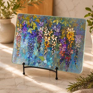 A rectangular glass cutting board with a stained-glass wisteria design stands upright on a black metal display stand on a white marble counter.