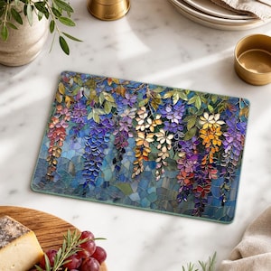 A rectangular glass cutting board with a stained-glass wisteria design is on a wooden surface, with a wedge of cheese and red grapes in the foreground.