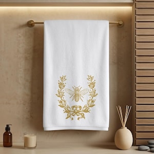 May include: White bath towel with a gold embroidered bee and laurel wreath design. The towel is hanging on a gold bar in a bathroom setting. The bee design is centered on the towel.