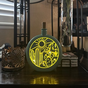 May include: A teal blue glass bottle with a cork top and a cut-out design of a desert scene with a sun, cactus, and mountains. The bottle is lit from inside, creating a warm glow.
