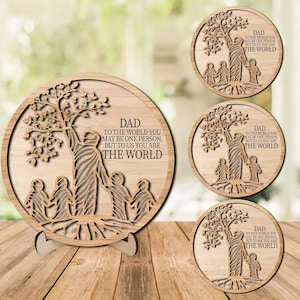 May include: Wooden round plaque with a laser-cut design featuring a tree, figures, and the text "DAD TO THE WORLD YOU MAY BE ONE PERSON, BUT TO US YOU ARE THE WORLD." Includes a stand. Several similar plaques are also shown.