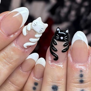 May include: Close-up of manicured nails with white and black cat designs. The nails have a natural base with white tips. One nail features a white cat, and another has a black cat, both in 3D relief.