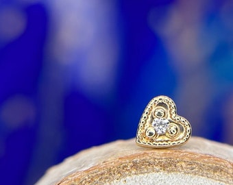 May include: A gold-colored heart-shaped stud earring with a small, clear gemstone in the center. The earring has a decorative border and is set against a blurred blue and white background.