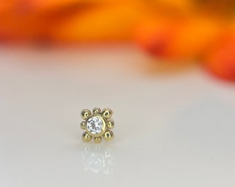 May include: A small, gold-toned nose ring with a clear gemstone in the center. The ring is surrounded by a circle of small, gold beads.
