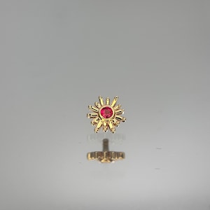 May include: A gold-colored stud earring with a sunburst design. The center features a round, red gemstone. The earring has a screw-on post. The design is symmetrical and detailed, with a polished finish.