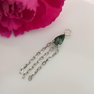 May include: A silver-toned pendant featuring a pear-shaped moss agate gemstone with a dark green, moss-like inclusion. The setting includes a small circular bail at the top and three dangling silver-toned metal chains of equal length.