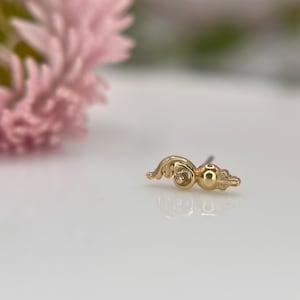 Puede incluir: Un pequeño pendiente dorado con un diseño ornamentado en forma de voluta. El pendiente presenta una pequeña bola y una forma de hoja. El fondo incluye una flor rosa borrosa y una superficie blanca.