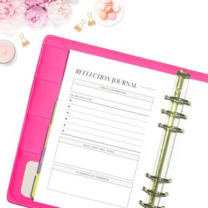 May include: A bright pink Reflection Journal with gold detailing. The journal is open to a page with prompts for daily reflection, including "Today's Affirmation" and "I'm grateful for...". The image also shows flowers, macarons, and gold paper clips.