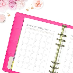 May include: A pink planner with a gold coil binding. The planner has a white page with the text "NO SPEND CHALLENGE" and a calendar grid with the days of the month numbered 1 through 30.