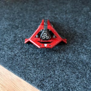 May include: A small, red, triangular object with a black central component and three circular openings. The object has a geometric design and appears to be made of metal or a similar material. The object is on a dark grey surface.