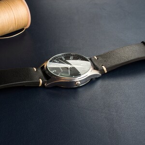 Puede incluir: Un reloj plateado con una esfera negra y agujas blancas. El reloj tiene una correa de cuero negra con costuras blancas.