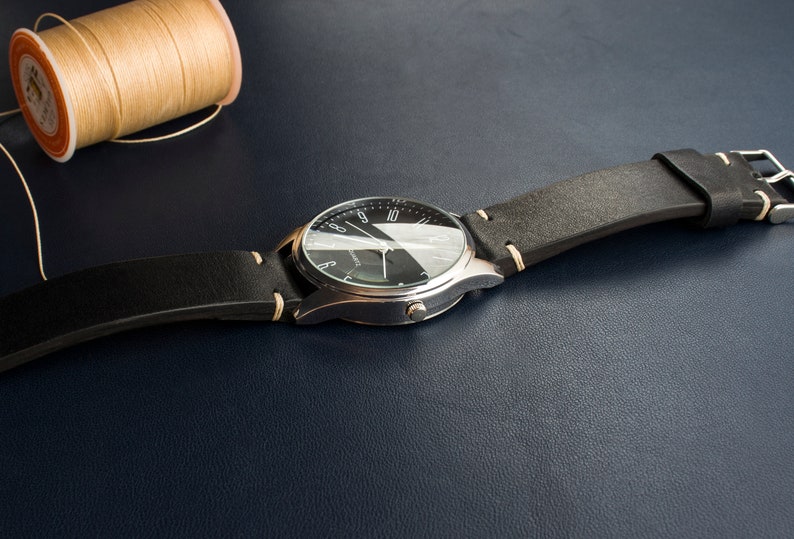Puede incluir: Un reloj plateado con una esfera negra y agujas blancas. El reloj tiene una correa de cuero negra con costuras blancas.