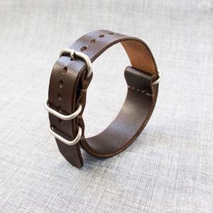 Puede incluir: Una correa de reloj de cuero marr&oacute;n con hebilla y trabillas de metal plateado.