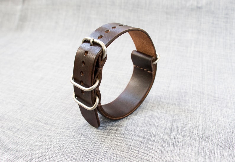 Puede incluir: Una correa de reloj de cuero marr&oacute;n con hebilla y trabillas de metal plateado.