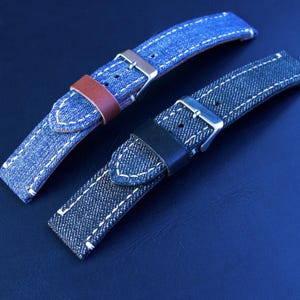 Puede incluir: Dos correas de reloj de mezclilla con costuras blancas y hebillas plateadas. Una correa tiene un acento de cuero marrón, la otra tiene un acento de cuero negro. Las correas se muestran sobre una superficie azul oscuro.