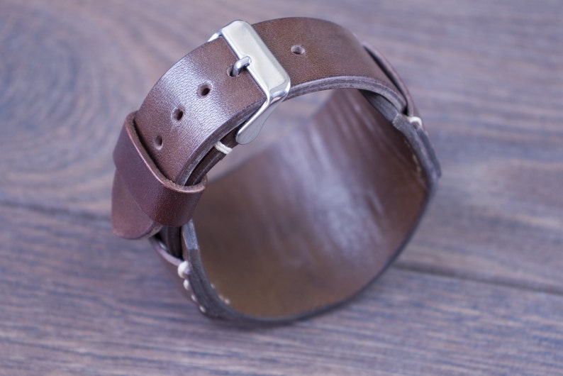 Puede incluir: Una correa de reloj de cuero marr&oacute;n con una hebilla plateada. La correa est&aacute; doblada por la mitad y se encuentra sobre una superficie de madera.